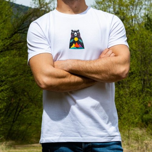 byala-teniska-s-mechka-planinar Man wearing a white unisex T-shirt with a small "Boho" bear design on the chest, featuring a colorful mountain landscape and a rising sun.
