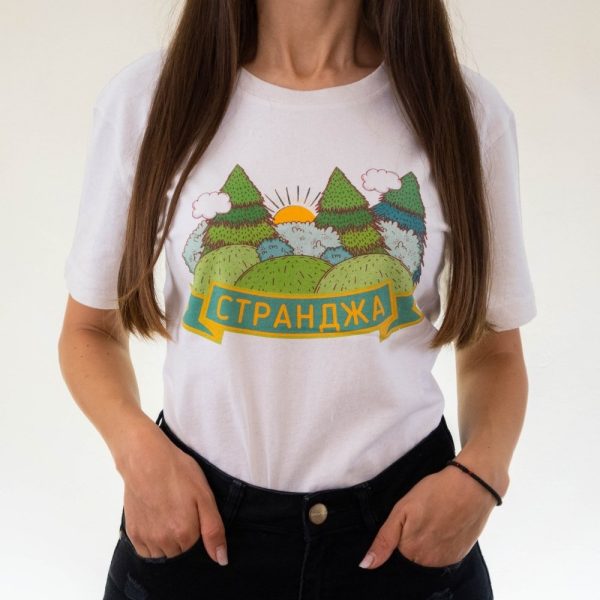 Woman wearing a white unisex T-shirt with a colorful illustration of mountains, trees, clouds, and the sun, featuring the word "Странджа" in Cyrillic.