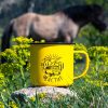 kanche-za-planina-kenche-camper-zhylto Yellow enamel camping mug with a camper van illustration and the word "Щастие" (Happiness), held by a hand with pink nail polish. Blurred train and green nature in the background.