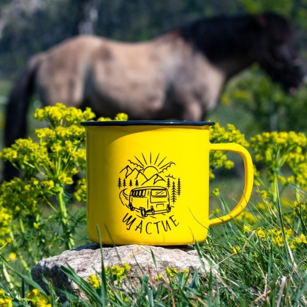 kanche-za-planina-kenche-camper-zhylto Yellow enamel camping mug with a camper van illustration and the word "Щастие" (Happiness), held by a hand with pink nail polish. Blurred train and green nature in the background.