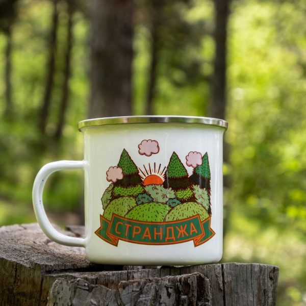 kanche-za-planina-kenche-metalno (10) White enamel camping mug featuring a colorful illustration of Strandzha Mountain – rolling green hills, sun rising behind peaks, and fluffy clouds. Placed on a wooden stump with a lush forest in the background.