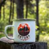 kanche-za-planina-kenche-metalno (11) White enamel camping mug with a colorful illustration of Vitosha Mountain – a stylized sunset over forested peaks. Placed on a wooden stump in a lush forest.