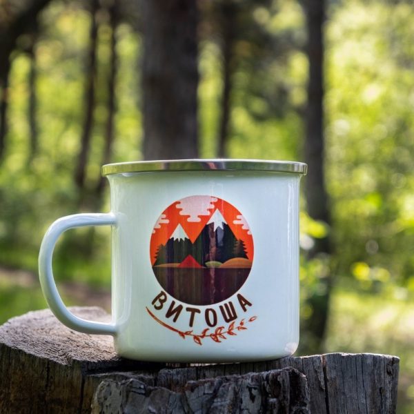 kanche-za-planina-kenche-metalno (11) White enamel camping mug with a colorful illustration of Vitosha Mountain – a stylized sunset over forested peaks. Placed on a wooden stump in a lush forest.