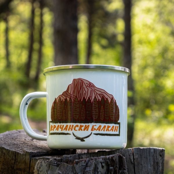 White enamel camping mug featuring an illustration of Vratsa Balkan’s rocky peaks and dense forests. Placed on a wooden stump in a lush green forest.