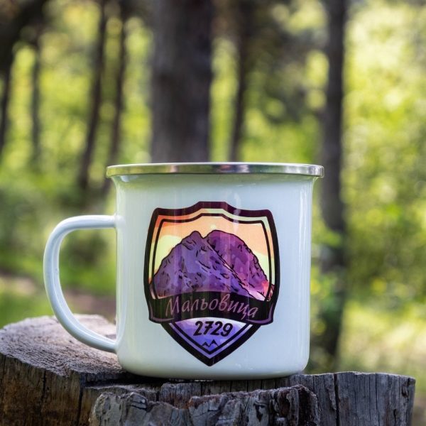 White enamel camping mug with a colorful design of Mount Malyovitsa (2729m), placed on a wooden stump in a forest background.
