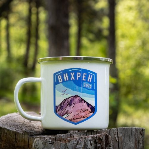 White enamel camping mug with a blue mountain shield design of Mount Vihren (2914m), placed on a wooden stump in a lush green forest.