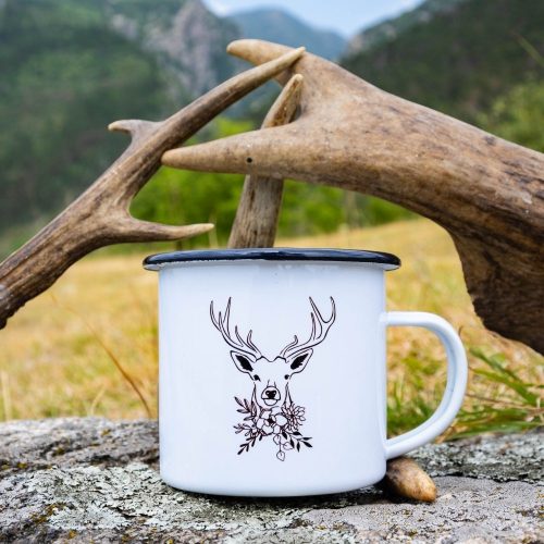 White enamel camping mug with a black rim, featuring a minimalist deer illustration with floral details. Placed on a rock with antlers in the background.
