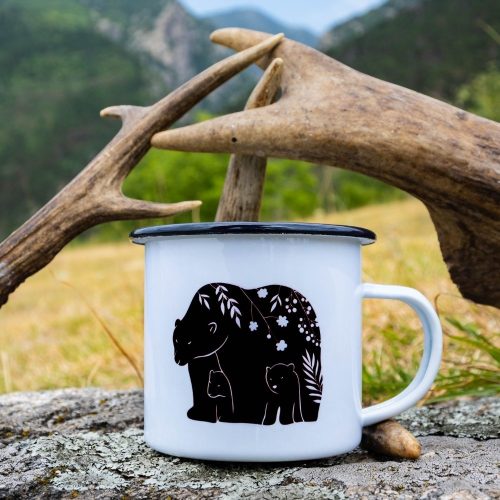 White enamel camping mug with a black rim, featuring a minimalist bear and cubs illustration with floral details. Placed on a rock with antlers in the background.