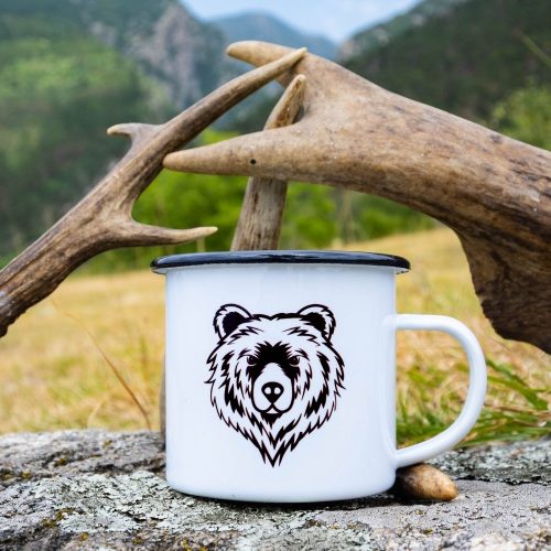 White enamel mug with a black rim, featuring a bold bear face illustration. Placed on a rock with antlers in the background.