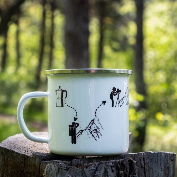 kanche-za-planina-kenche-metalno (3) White enamel mug with a minimalist adventure-themed design, featuring a coffee maker, a hiker, a mountain, and a photographer. Placed on a wooden stump with a green forest background.
