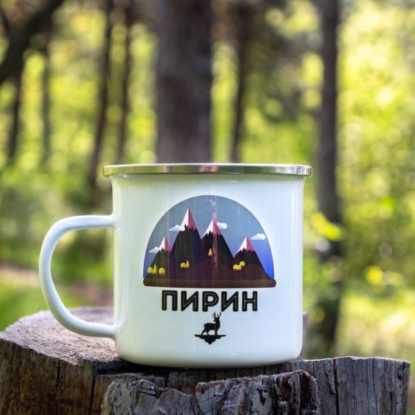 White enamel camping mug with a stylized design of the Pirin Mountains, featuring alpine peaks, wild goats, and a deer silhouette. Placed on a wooden stump with a green forest background.
