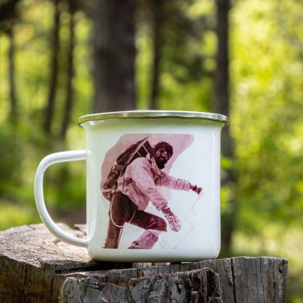 kanche-za-planina-kenche-metalno (5) White enamel camping mug featuring a vintage alpinist photograph, placed on a wooden stump with a lush green forest in the background.