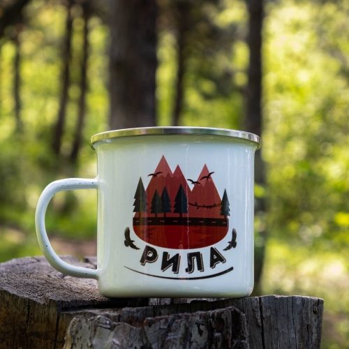White enamel camping mug with a stylized design of the Rila Mountains, featuring peaks, pine trees, and flying birds. Placed on a wooden stump with a lush green forest in the background.