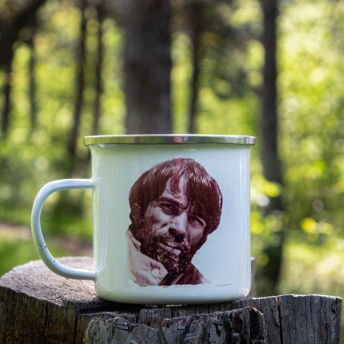 kanche-za-planina-kenche-metalno (7) White enamel camping mug featuring a vintage portrait of a mountaineer, placed on a wooden stump with a lush green forest in the background.