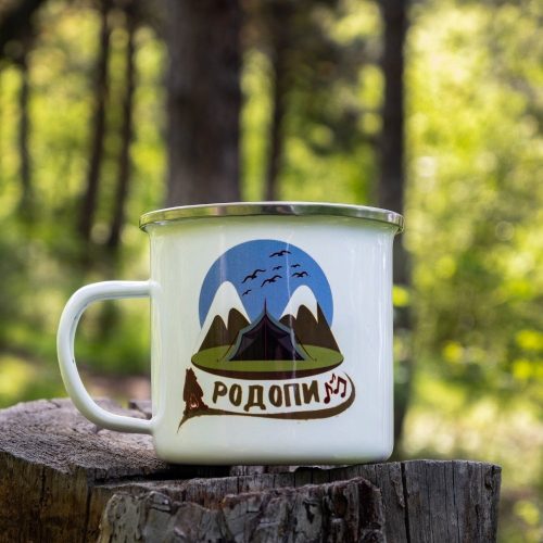 White enamel camping mug with a design of the Rhodope Mountains, featuring a tent, snowy peaks, and birds in the sky. Placed on a wooden stump with a green forest in the background.