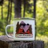 White enamel camping mug with a colorful design of the Balkan Mountains, featuring snowy peaks, a mountain hut, and trees. Placed on a wooden stump with a lush green forest in the background.