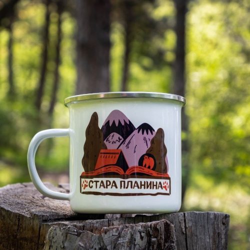 kanche-za-planina-kenche-metalno (9) White enamel camping mug with a colorful design of the Balkan Mountains, featuring snowy peaks, a mountain hut, and trees. Placed on a wooden stump with a lush green forest in the background.