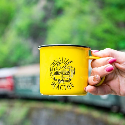 kanche-za-planina-kenche-shtastie Yellow enamel camping mug with a camper van illustration and the word "Щастие" (Happiness), held by a hand with pink nail polish. Blurred train and green nature in the background.