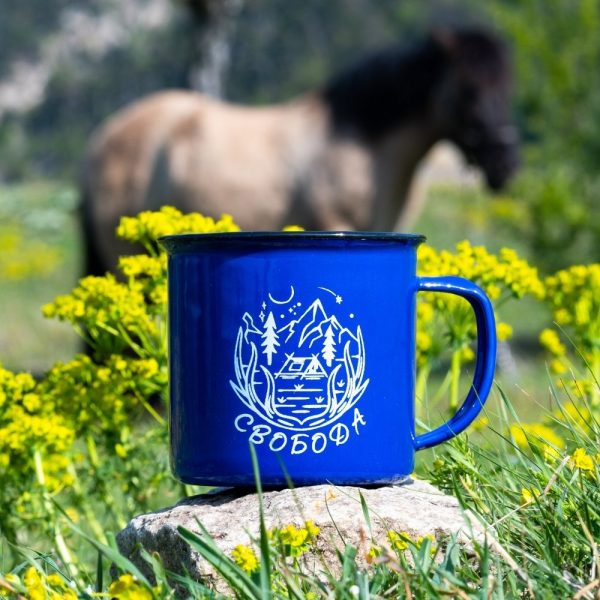 kanche-za-planina-kenche-sinio-svoboda Blue enamel camping mug with a mountain cabin illustration and the word "Свобода" (Freedom), placed on a rock with yellow wildflowers and a blurred horse in the background.