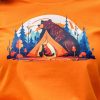 Close-up of an orange T-shirt featuring a detailed illustration of a bear and cub near a glowing tent in a forest setting.