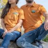 polo-teniska-s-yaka-oranzheva-brodirana A man and a woman wearing matching orange polo shirts with a small embroidered bear and mountain landscape design on the chest.