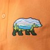 polo-teniska-s-yaka-oranzheva-s-broderia Close-up of an embroidered bear design on an orange polo T-shirt, featuring a mountain landscape inside the bear silhouette.