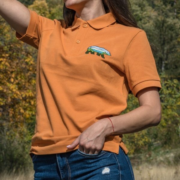 polo-teniska-s-yaka-oranzheva-zhenska Woman wearing an orange polo T-shirt with an embroidered bear and mountain landscape design.