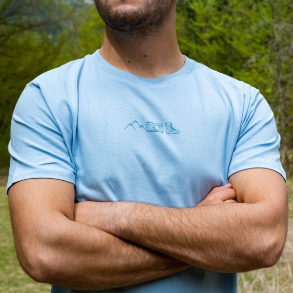 sinia-teniska-myzh-planina Man wearing a light blue unisex T-shirt with a minimalist design featuring mountain peaks, a camera, and a hiking boot.