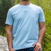 svetlo-sinia-teniska-za-planinar Man wearing a light blue unisex T-shirt with a minimalist "Simple" design featuring mountain peaks, a camera, and a hiking boot.