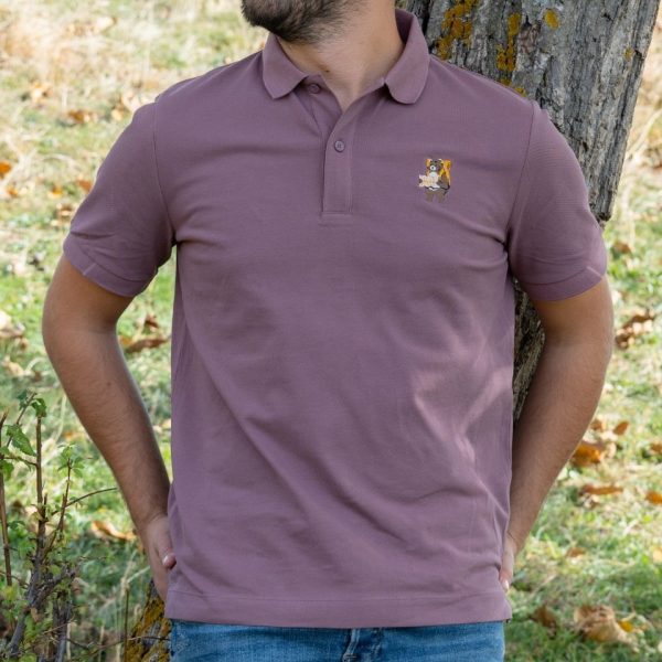 Man wearing a muted mauve polo shirt with an embroidered design of a bear holding a map, standing against a tree in a natural setting.
