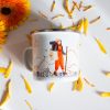Podarak-za-momiche-zhena-planinarka (5) White enamel camping mug featuring an illustration of a female hiker with a backpack and trekking pole, walking through a mountain landscape, surrounded by yellow flower petals.