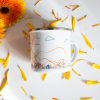 Podarak-za-momiche-zhena-planinarka (6) White enamel camping mug featuring a minimalist mountain landscape with orange contour lines, clouds, birds, and wildflowers, surrounded by yellow flower petals.