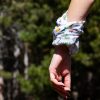 bandana-za-planina-explorebg Close-up of a woman's hand wearing a multifunctional white buff as a wristband, featuring hiking-themed illustrations and Bulgarian mountain names.