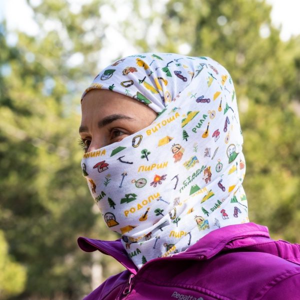 bandana-za-planina-maska-damska Woman wearing a white multifunctional buff as a full-face cover and headscarf, featuring Bulgarian mountain names and hiking-themed illustrations.