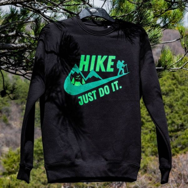 Black fleece sweatshirt with "HIKE – JUST DO IT" print, hanging on a tree branch in a natural setting.
