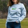 A woman wearing a light blue sweatshirt with an embroidered bear featuring a mountain landscape design, standing in a natural outdoor setting.