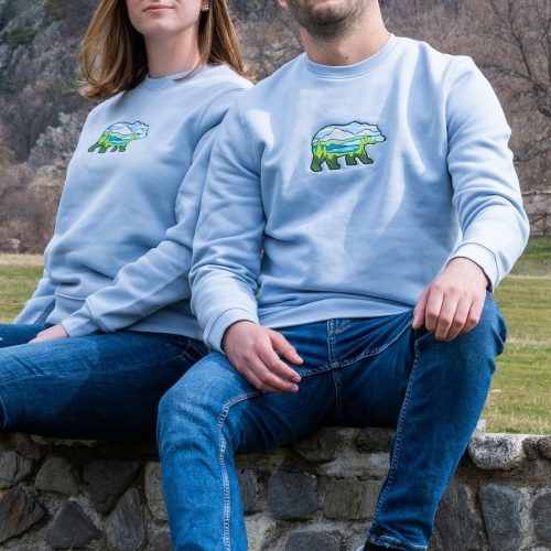 bluza-s-broderia-sinia-mechka-explorebg Matching light blue sweatshirts with an embroidered bear and mountain landscape design, worn by a man and a woman outdoors.