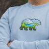 Close-up of a light blue sweatshirt featuring an embroidered bear with a mountain landscape inside.