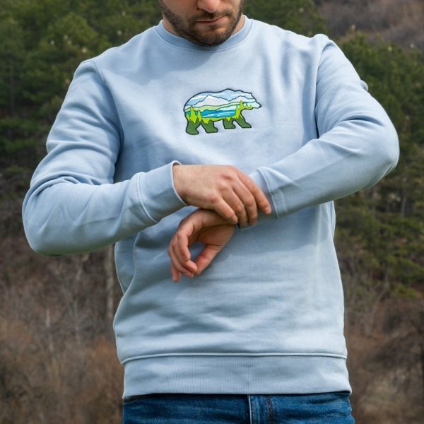 A man wearing a light blue sweatshirt with an embroidered bear featuring a mountain landscape, adjusting his sleeve in an outdoor setting.