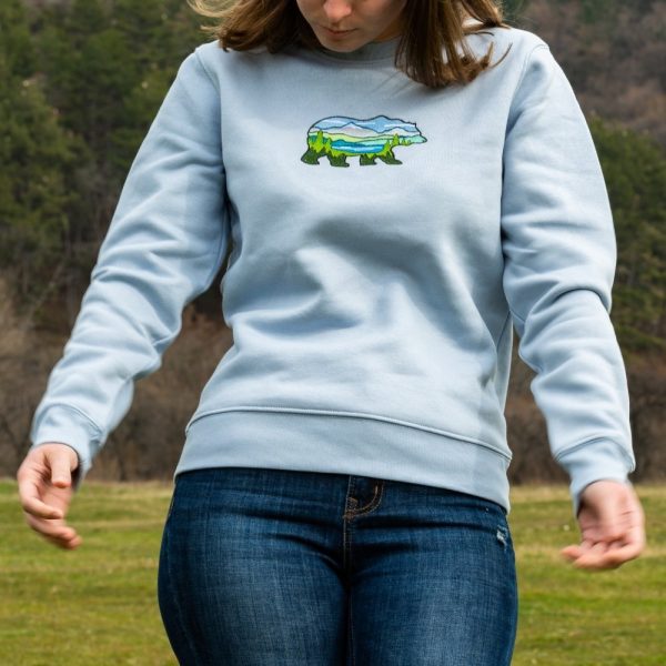 A woman wearing a light blue sweatshirt with an embroidered bear featuring a mountain landscape, adjusting her sleeves outdoors.