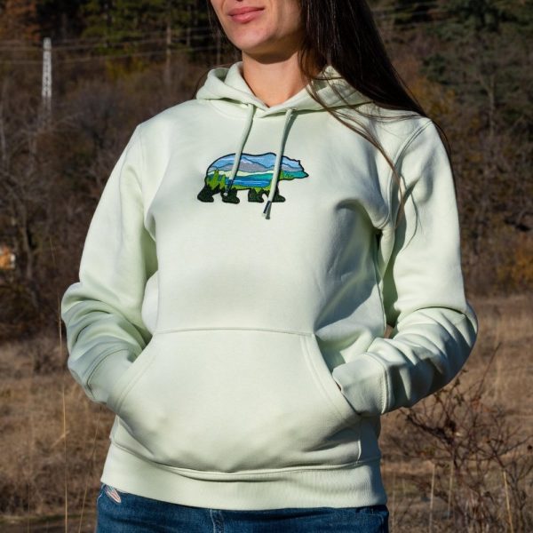 Light green hoodie with an embroidered bear featuring a scenic mountain landscape, worn by a woman outdoors.