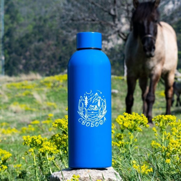 Blue stainless steel bottle (800 ml) with an illustration of a cabin in the mountains and the word "Свобода" (Freedom), placed on a rock in a natural setting with a horse in the background.