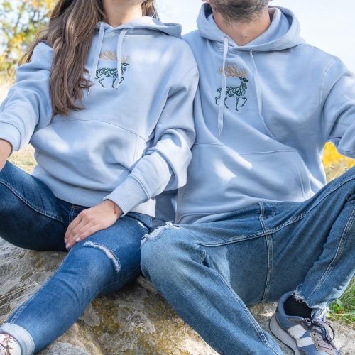 hoodie-s-kachulka-sinio Light blue hoodies with embroidered moose design, worn by a couple sitting outdoors.
