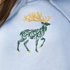 hoodie-s-kachulka-sinio-elen Close-up of an embroidered deer design on a light blue oversized hoodie, featuring intricate green and golden details.