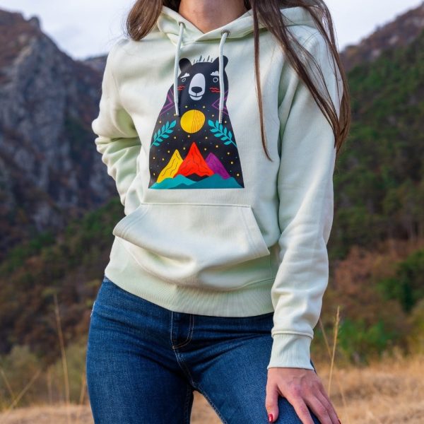 hoodie-sweatshirt-zelen-cvyat Light green hoodie featuring a colorful bear and mountain print, worn by a woman outdoors.