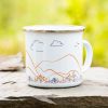 White enamel camping mug featuring a minimalist landscape illustration with rolling mountains, clouds, birds, and wild grasses in warm earthy tones.