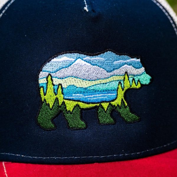 Close-up of an embroidered bear landscape design on a navy blue trucker hat with a red brim.