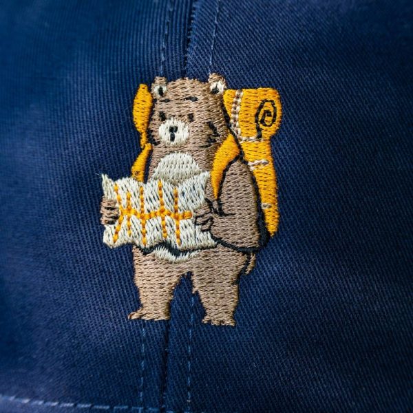 Close-up of embroidered bear with a backpack and a map on a navy blue cap.
