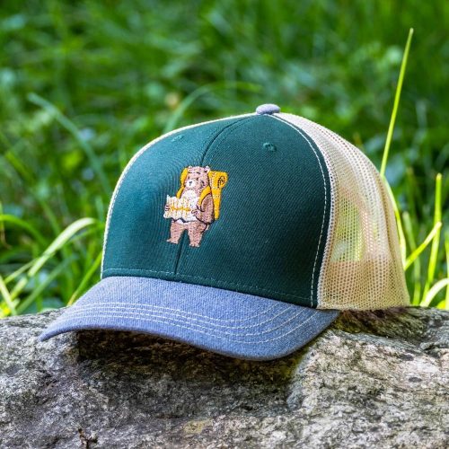 shapka-s-kozirka-i-mrezha-siva Green and denim cap with embroidered bear carrying a backpack and a map, placed on a rock in nature.