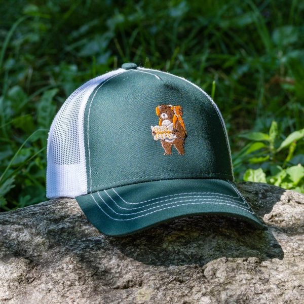 Green and white trucker cap with embroidered hiking bear design, placed on a rock outdoors.